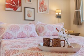 Uma cama bem arrumada com roupa de cama florida, um tabuleiro com duas canecas e um vaso com orquídeas brancas.