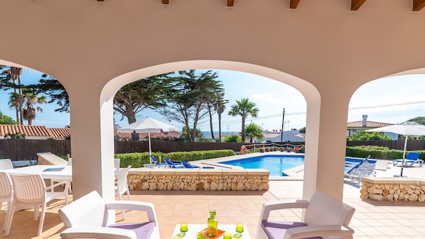 Un espace extérieur aménagé, abrité et doté de mobilier en osier blanc et de coussins violets, offrant une vue sur une piscine et un jardin paysager.