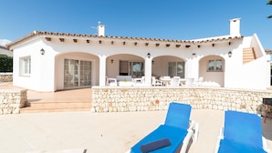 Un bâtiment blanc avec un toit en tuiles, un mur en pierre et un patio aménagé avec des chaises longues.