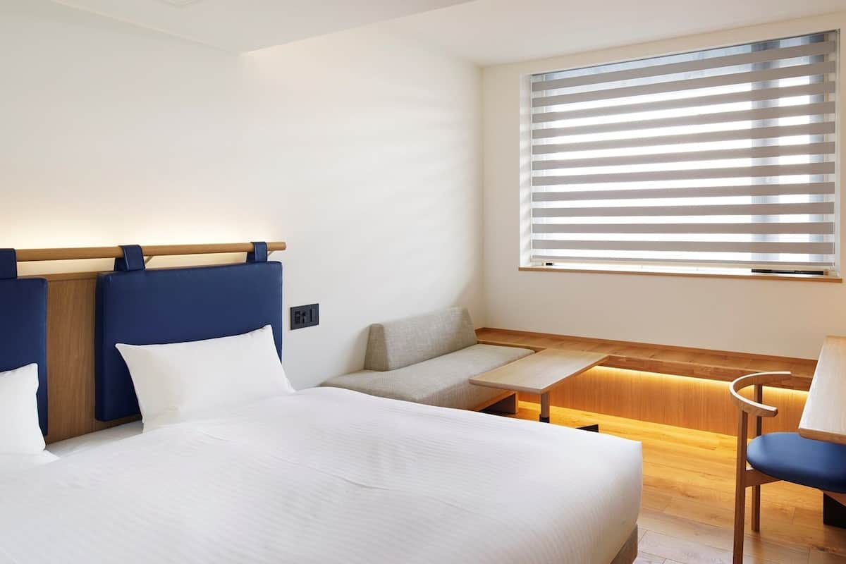 Une chambre d’hôtel comprenant un lit, un bureau, une chaise, un canapé et une fenêtre avec des stores.