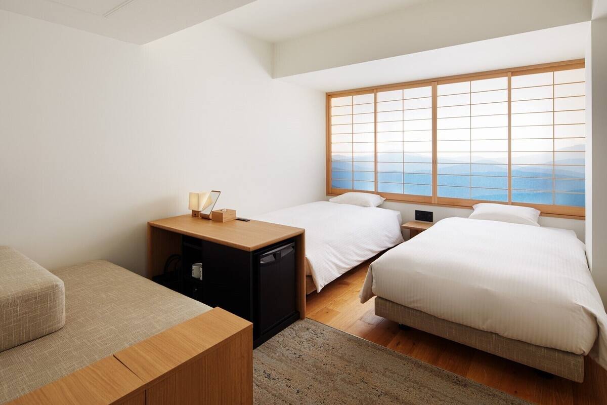 Une chambre d’hôtel avec deux lits, un bureau et une vue sur les montagnes, visible par une grande fenêtre.