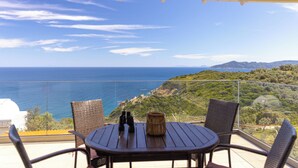 A balcony with wicker chairs, a round table, and a view of the sea and hills.