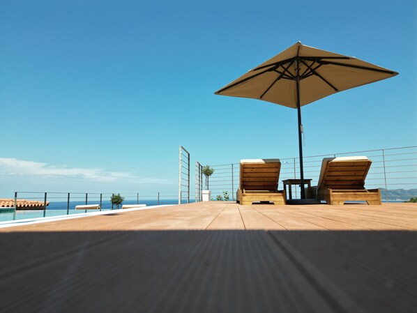 A wooden deck with two lounge chairs and a large umbrella, overlooking a body of water.