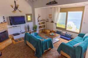 A living room with a fireplace, a television, a sofa set, a coffee table, and a view of the sea.