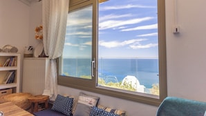 A room with a large window overlooking the sea, a wooden table, a sofa with patterned cushions, and a white radiator.
