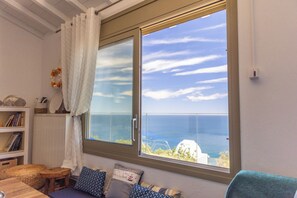 Een kamer met een groot raam met uitzicht op zee, een houten tafel, een bank met opvallende kussens en een witte radiator.