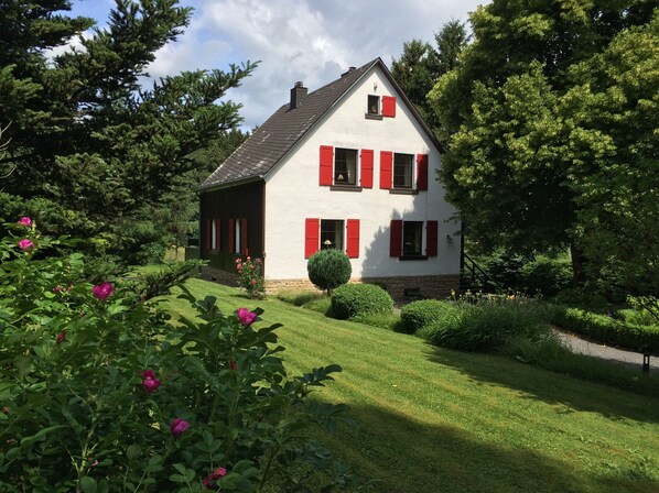 Charming single-story house with a steeply pitched roof and symmetrical facade, surrounded by lush greenery and well-maintained lawns.