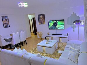 Un soggiorno moderno con un divano bianco, un tavolino da caffè e una televisione a schermo piatto. Si vedono quadri appesi alle pareti e un lampadario decorativo.