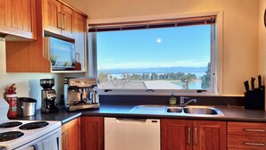 A kitchen with wooden cabinets, a stove, a coffee maker, and a view of a lake and mountains through a window. - Kimberley Three - Nelson Waterfront Apartment - Free Cancellation Available Here (Nelson)