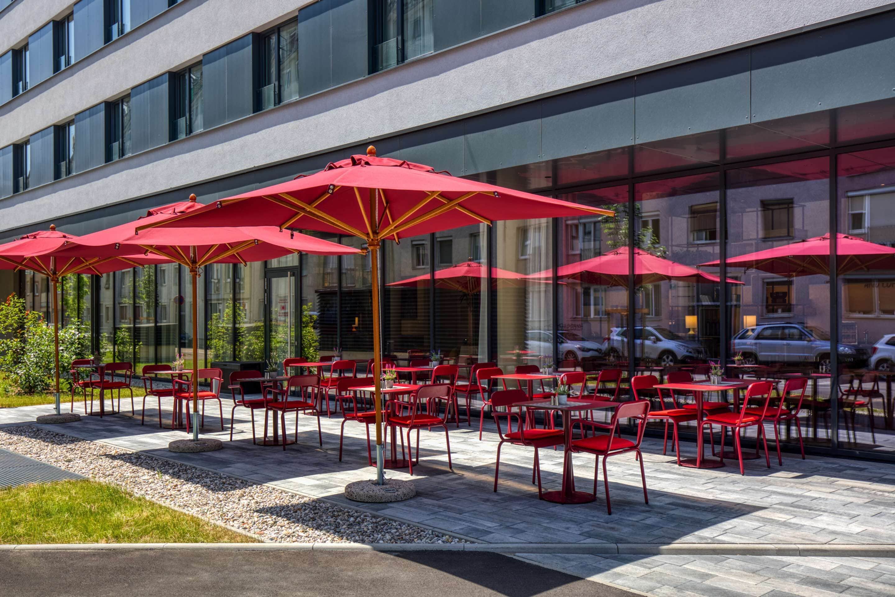 An outdoor seating area with red chairs and umbrellas, a modern building with large windows, and a paved patio.