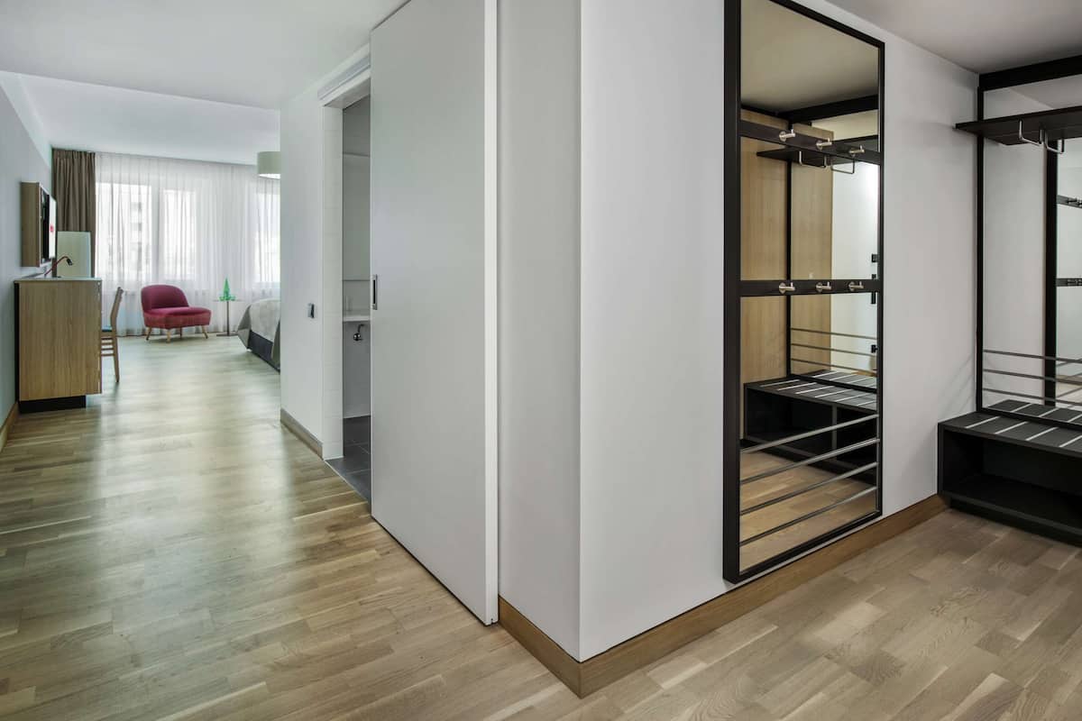 Una habitación de hotel moderna con piso de madera, una silla roja, una cama, un armario con espejo y una mesita de noche con lámpara.
