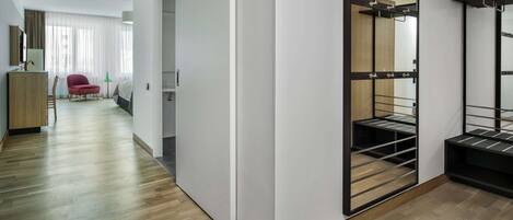 Ein modernes Hotelzimmer mit Holzboden, einem roten Sessel, einem Bett, einem Schrank mit Spiegel und einem kleinen Tisch mit Lampe.
