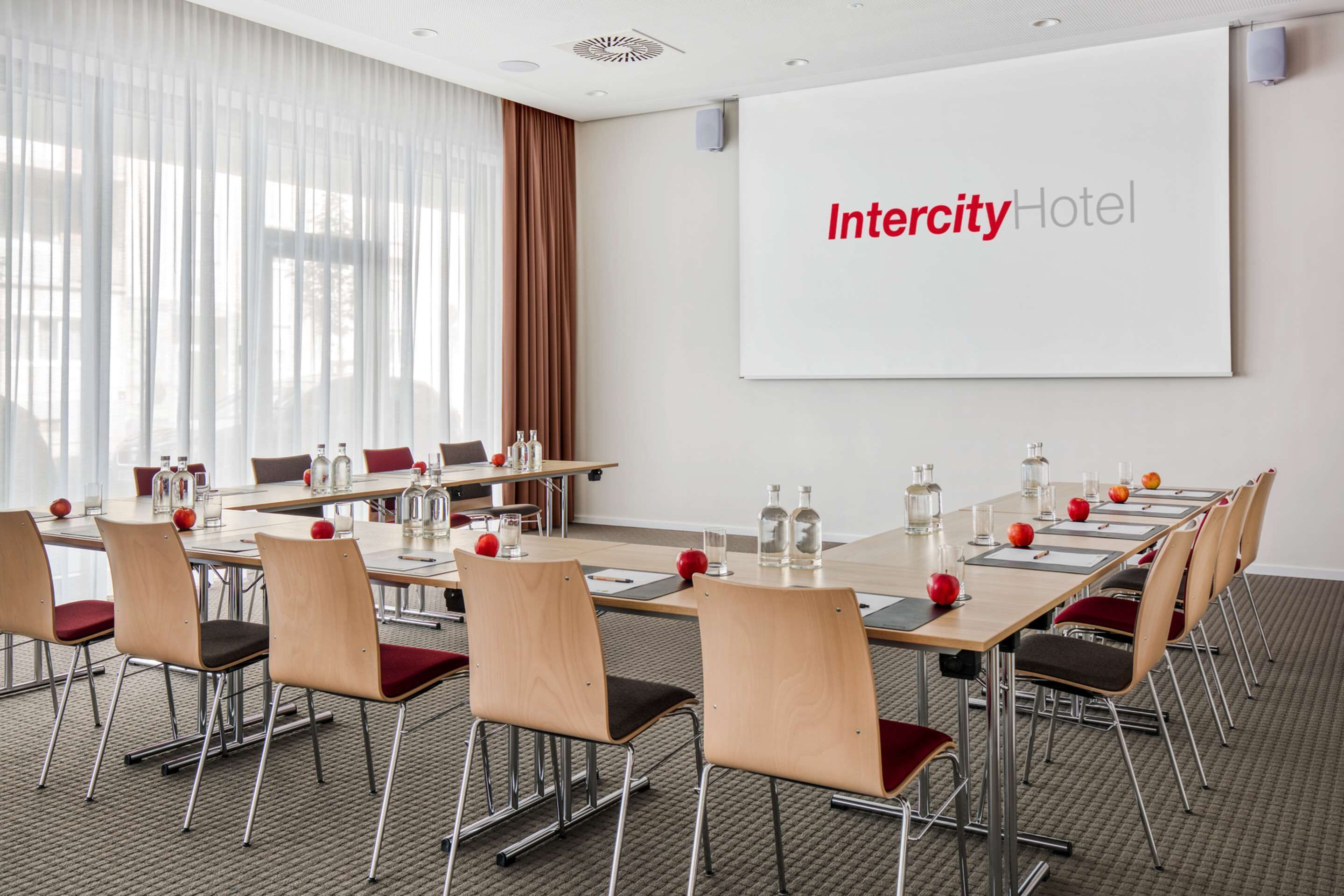 Egy konferenciaterem, ahol egy nagy képernyőn az „Intercity Hotel” felirat látható, és egy hosszú asztal áll, székekkel és ásványvizes palackokkal.