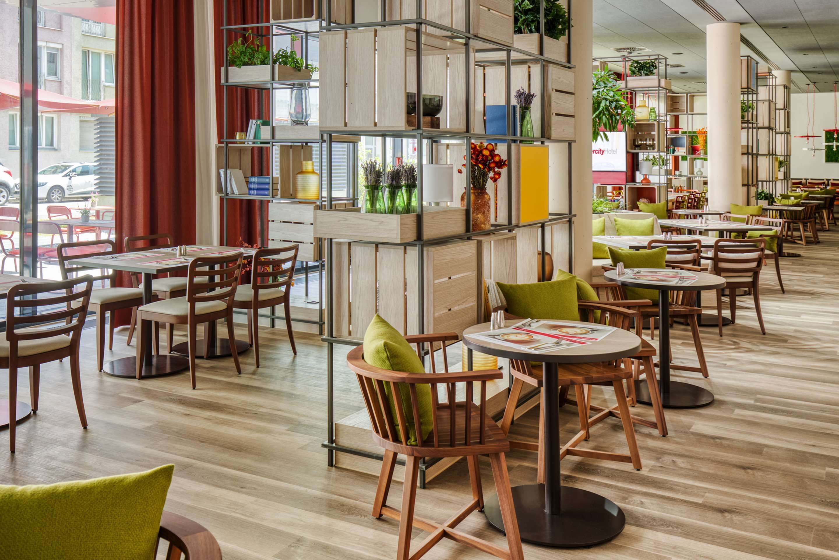 A modern cafe with wooden furniture, green cushions, and a variety of plants on shelves.