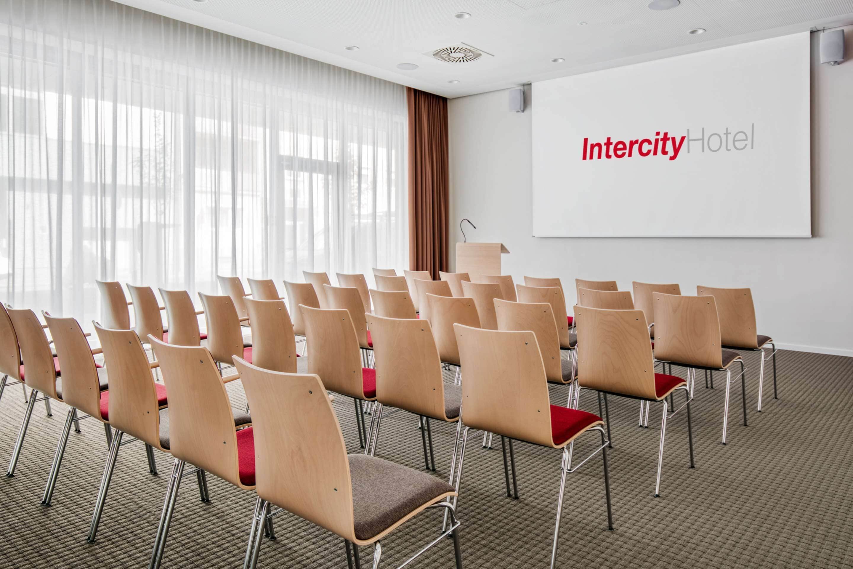 Uma sala de conferências com fileiras de cadeiras, uma tela de projetor e uma janela grande com cortinas.