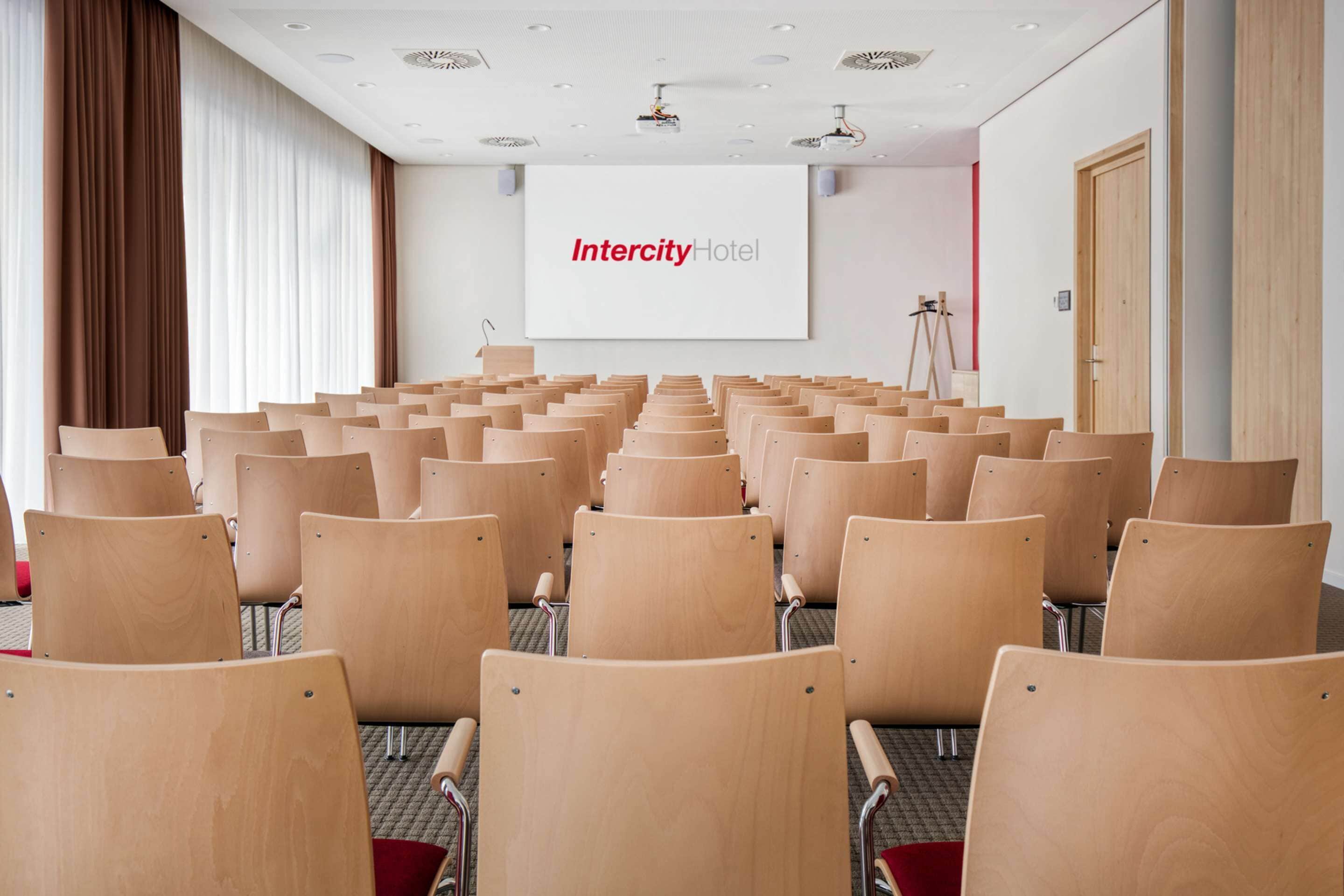 Un salón de conferencias con filas de sillas de madera, una pantalla grande que muestra "Intercity Hotel" y un proyector sobre un trípode.