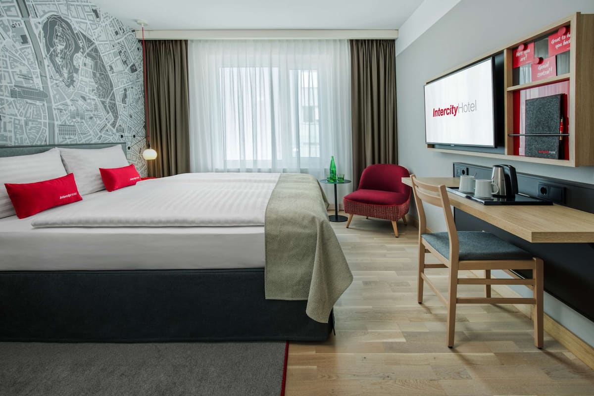 Habitación de hotel moderna con una cama grande, un escritorio, una silla y una pantalla de televisión mostrando el nombre del hotel.