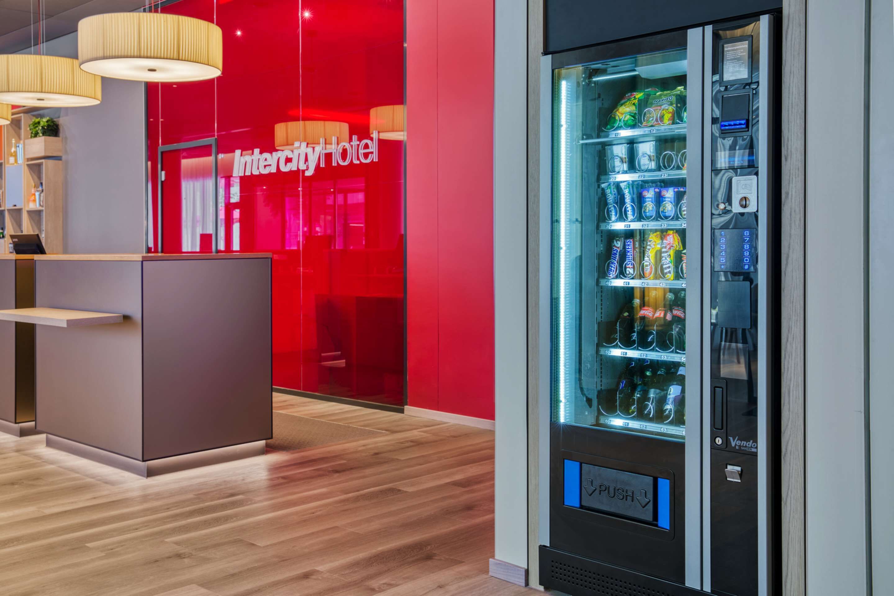 Een moderne hotellobby met een automaat, houten vloeren en een rode accentmuur.