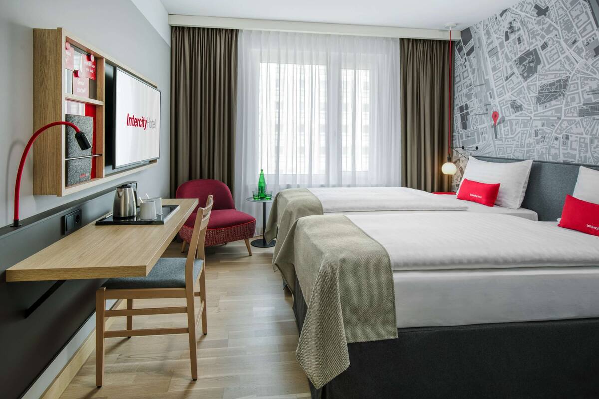 Habitación de hotel con escritorio, dos camas, una silla roja y un mural de mapa.
