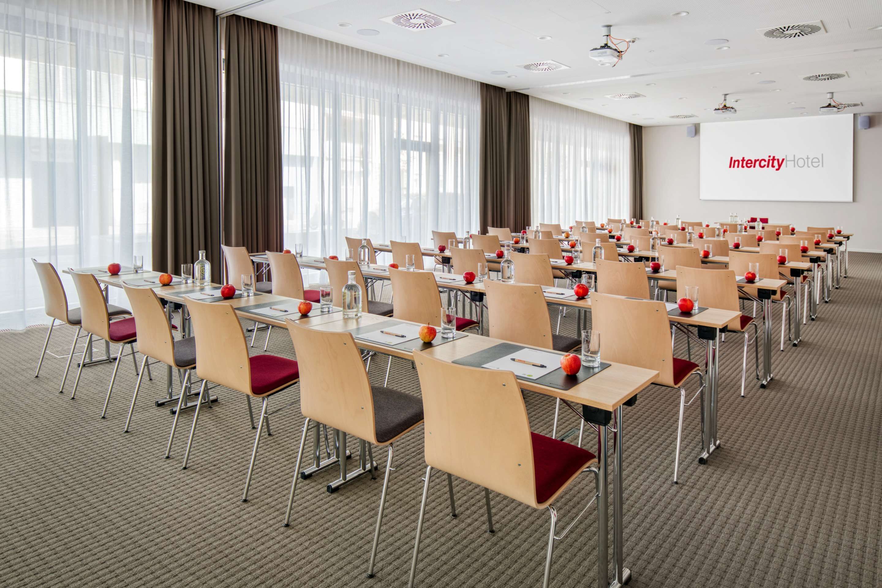 Un salón de conferencias con filas de mesas y sillas, una pantalla de proyector con el texto 'Intercity Hotel' y grandes ventanales con cortinas.
