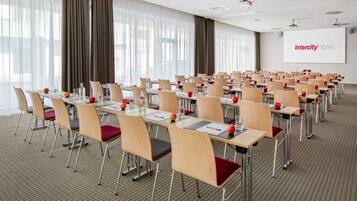 Un salón de conferencias con filas de mesas y sillas, una pantalla de proyector con el texto 'Intercity Hotel' y grandes ventanales con cortinas.