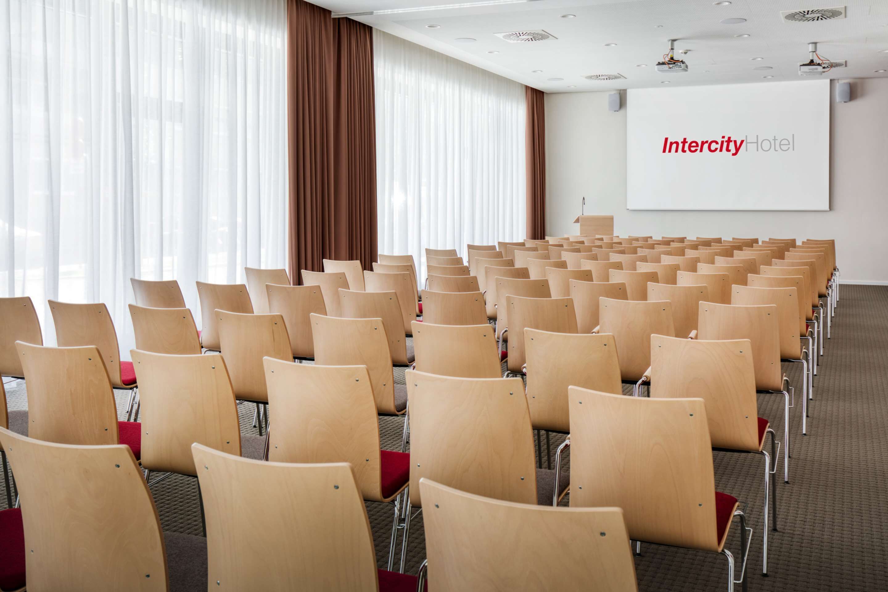 Un salón de conferencias con filas de sillas de madera y una pantalla grande que muestra 