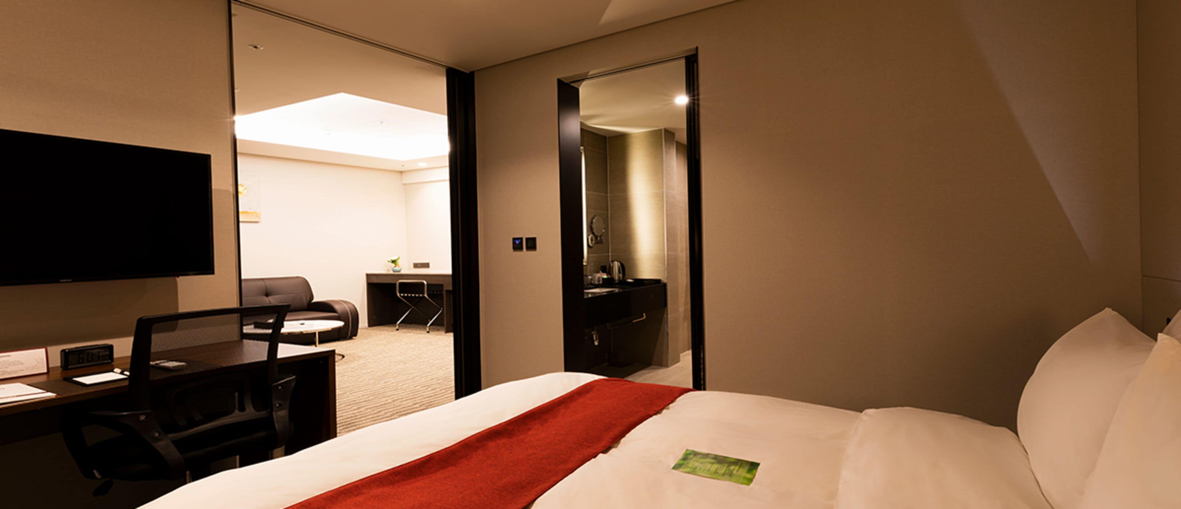 A hotel room with a bed, a desk with a chair, a television, and a bathroom visible through an open door.