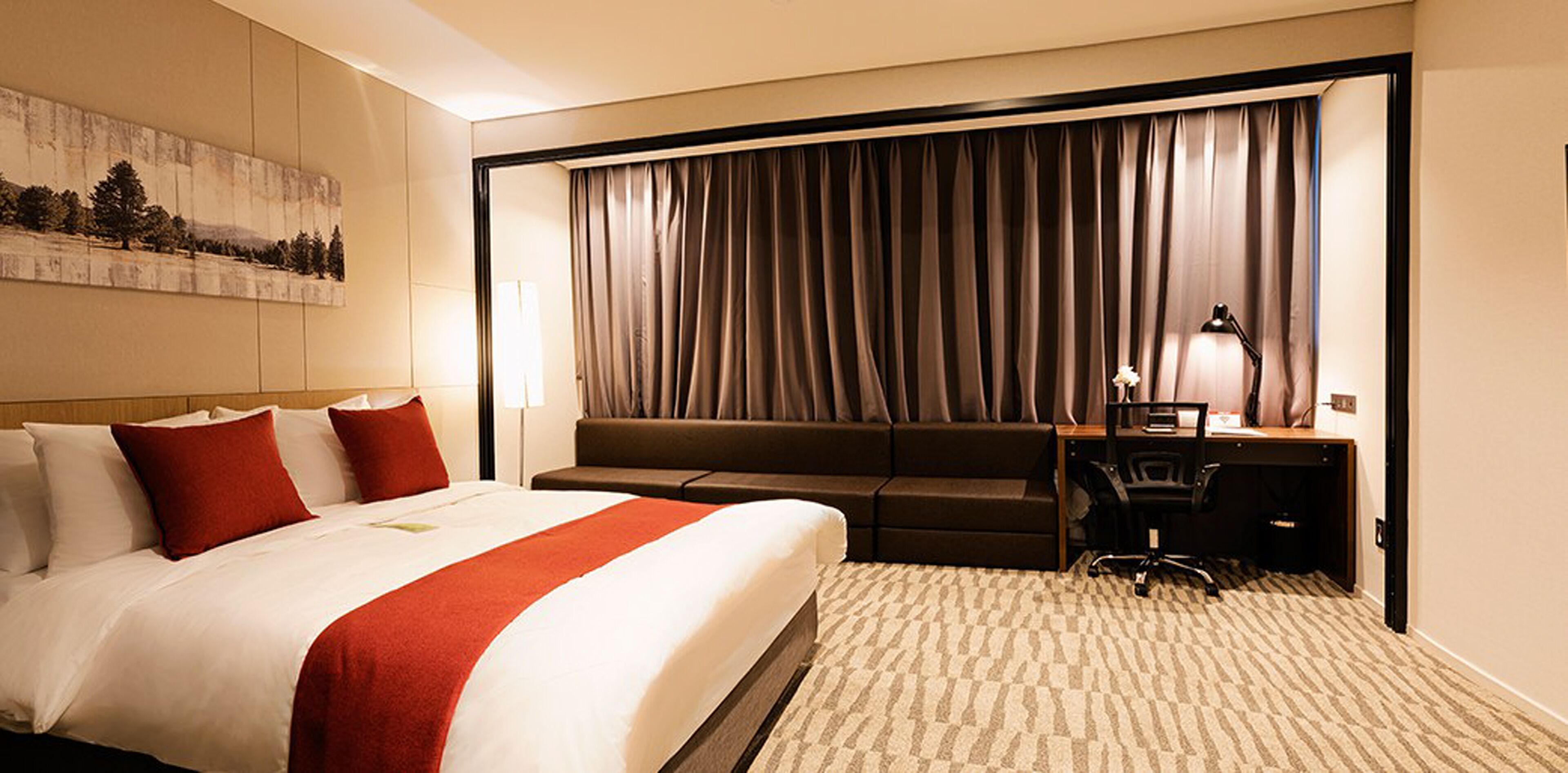 A hotel room with a large bed, a sofa, a desk, and a chair. There is a painting on the wall and a window with curtains.