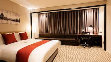 A hotel room with a large bed, a sofa, a desk, and a chair. There is a painting on the wall and a window with curtains.