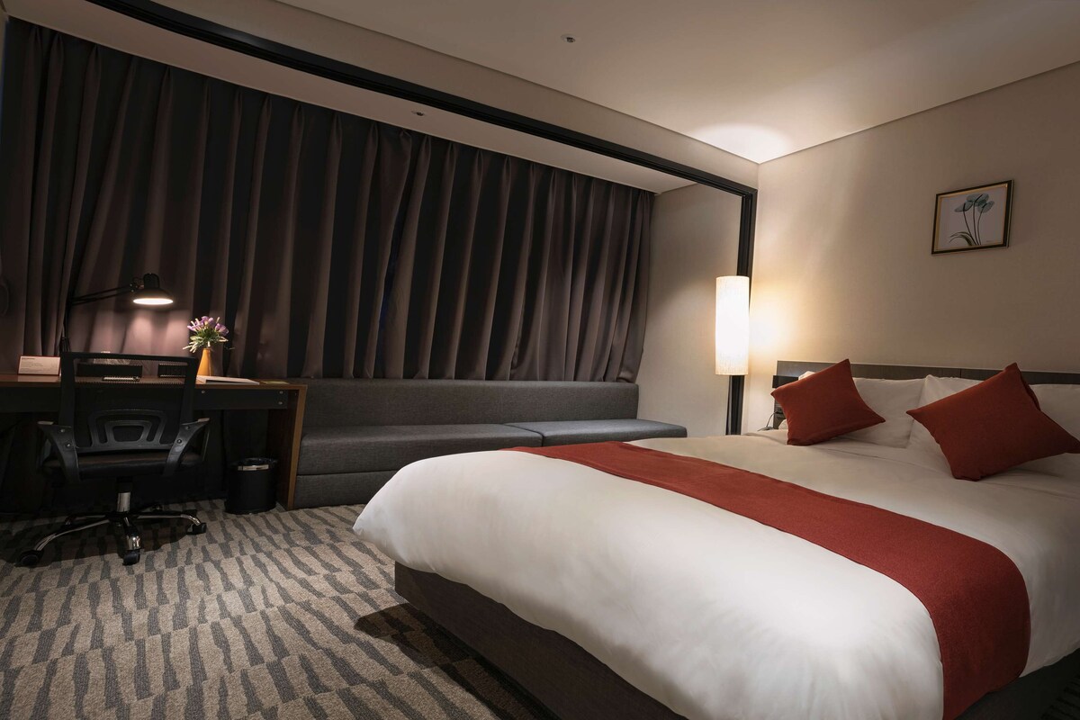A hotel room with a large bed, a desk, a chair, a sofa, and a lamp.