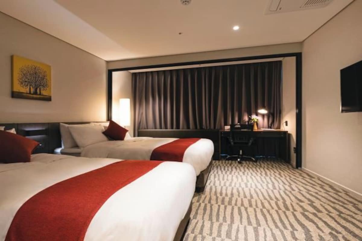 A hotel room with two beds, a desk, a chair, a TV, and a window with curtains.