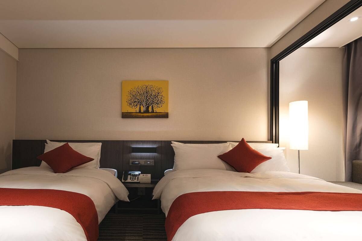 Ein Hotelzimmer mit zwei Betten, einer Bedienfeld, einem Baumgemälde und einer Lampe.