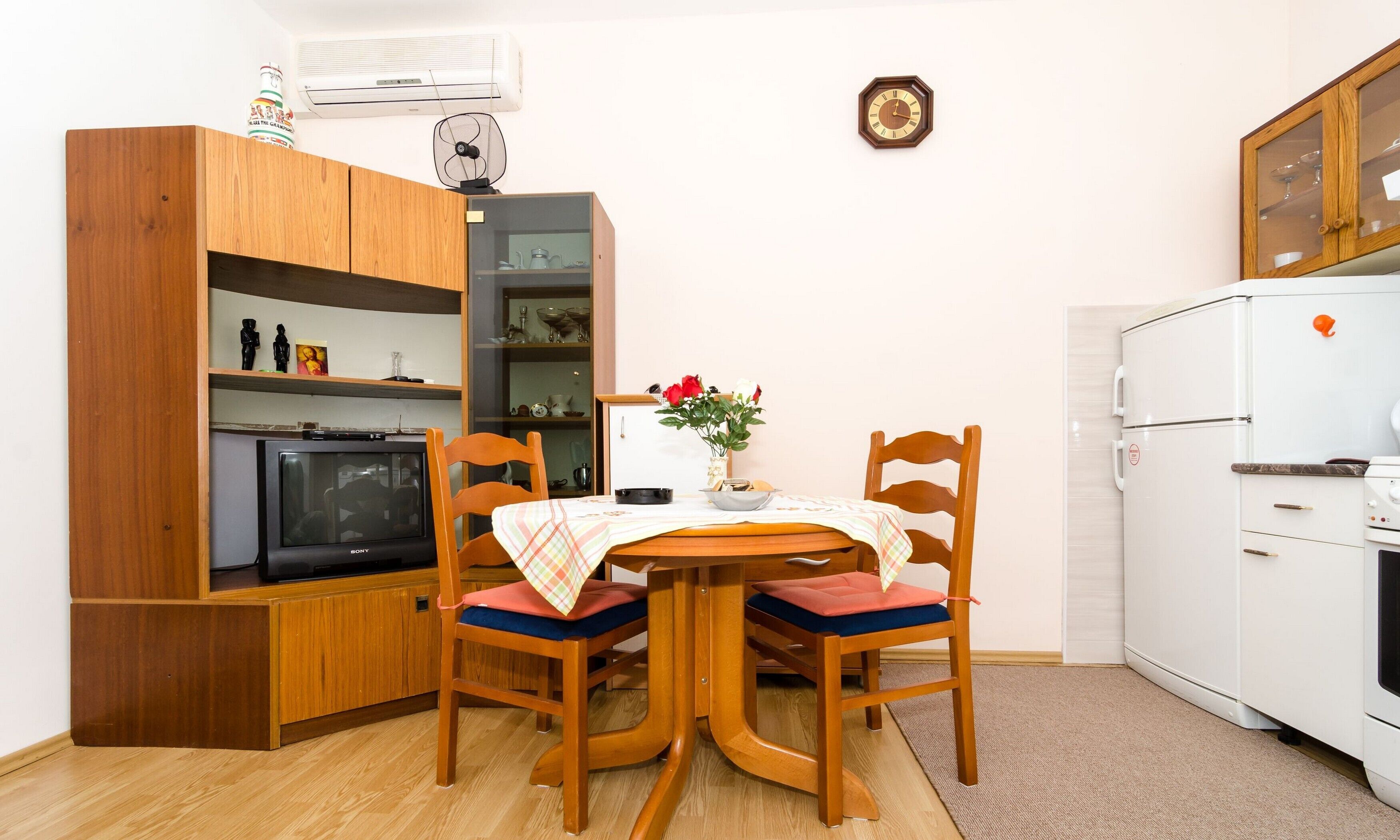 A compact kitchen with wooden cabinets, a dining table with chairs, a small refrigerator, and a wall-mounted clock.