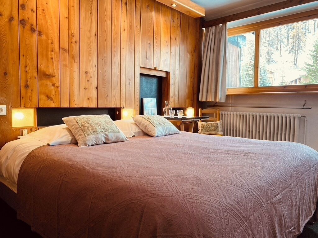 A bedroom with a large bed, wooden panel walls, a window with curtains, and a nightstand with a lamp.