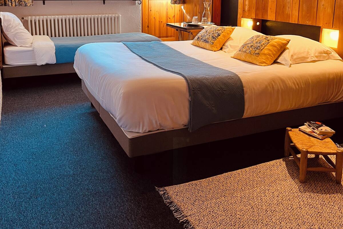 A bedroom with two beds, a wooden wall, a window with curtains, a radiator, and a bedside table with a lamp.