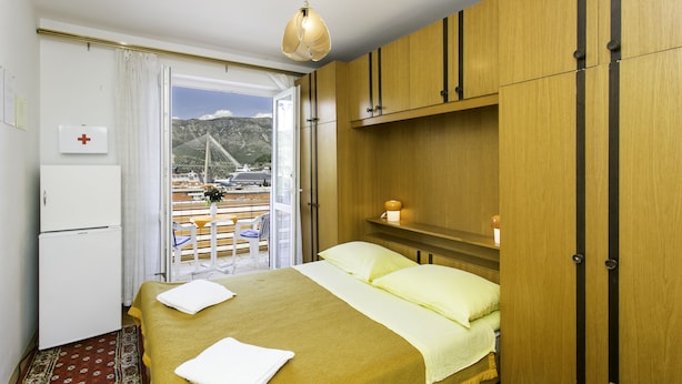Habitación de hotel con cama, una mesita, un armario, una mesita de noche con lámpara, una ventana con vista a un puente y barcos, y una puerta con un letrero de cruz roja.