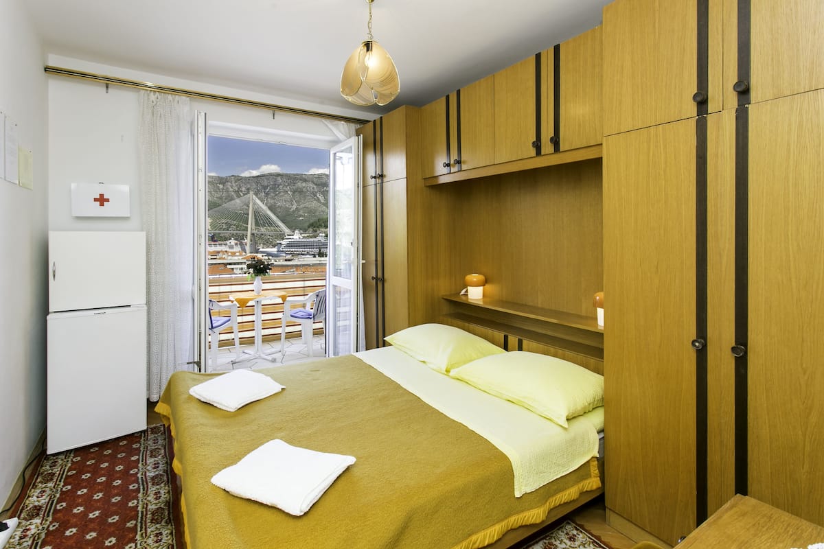 Une chambre d’hôtel avec un lit, une petite table, une armoire, une table de chevet avec une lampe, une fenêtre donnant sur un pont et des bateaux, et une porte avec un panneau représentant une croix rouge.