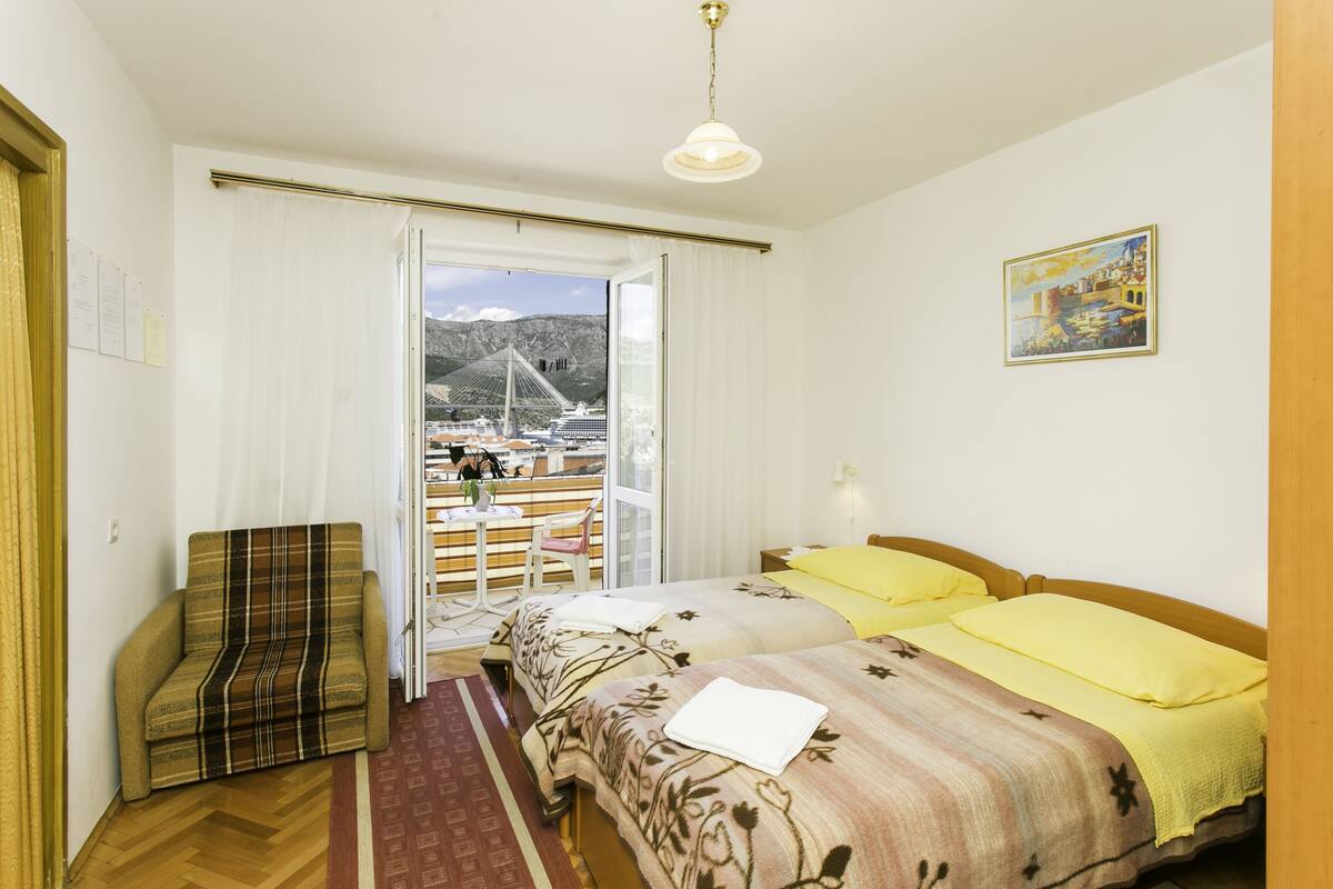 Une chambre d’hôtel avec un lit, une chaise, une fenêtre donnant sur un pont et des montagnes, et un tableau accroché au mur.