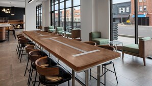 A modern bar area with a long wooden counter, multiple bar stools, and large windows offering views of the street. - Courtyard by Marriott Lansing Downtown (Lansing)