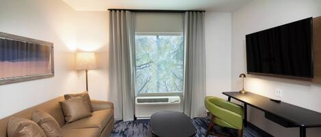 A hotel room with a sofa, a small table, a green chair, a TV, and a window with curtains.