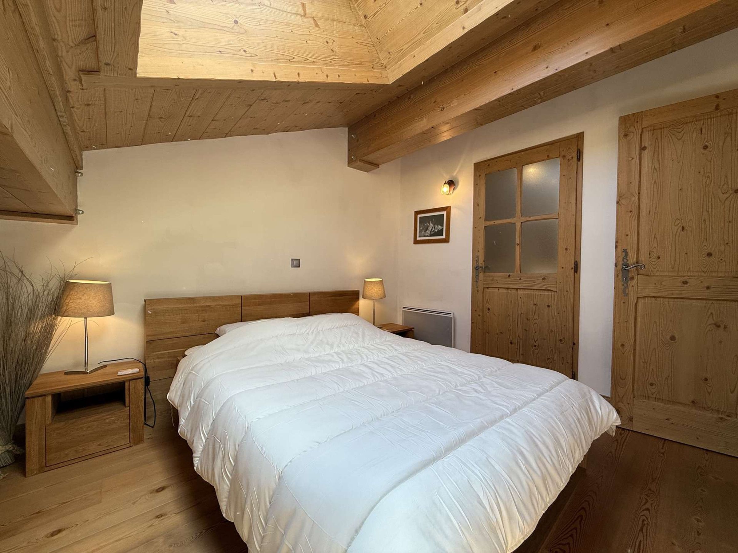 Un dormitorio con techo de madera, una cama con ropa de cama blanca, lámparas de noche, una mesita de noche, un radiador y puertas de madera.