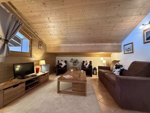 A cozy living room with a wooden ceiling, a television, a coffee table, a sofa, and a window with curtains.