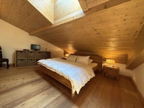 A bedroom with a sloped ceiling, wooden flooring, a bed with white bedding, a television on a wooden cabinet, and a nightstand with a lamp.