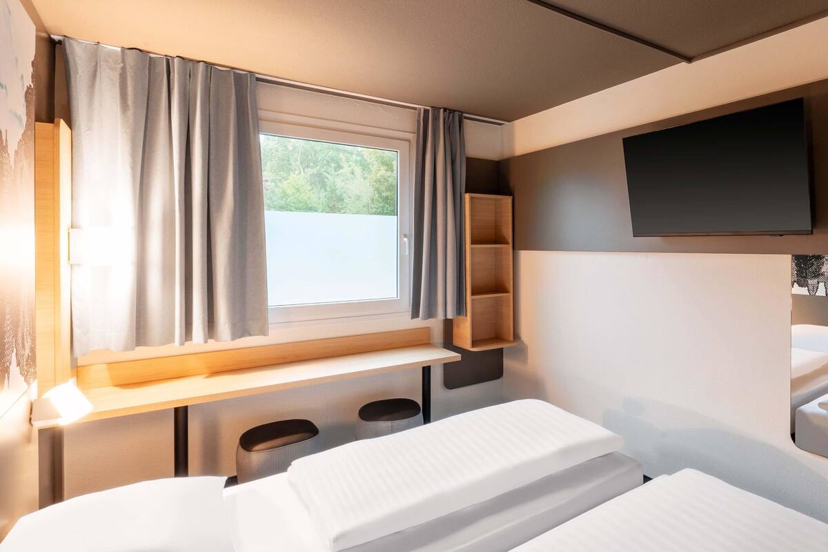 Een kleine slaapkamer met een enkel bed, een raam met gordijnen, een tv aan de muur en een klein plankje.