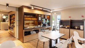 Un café moderne avec un comptoir où sont exposés des plats et des boissons, des chaises blanches disposées autour d’une table et une vue sur l’extérieur.