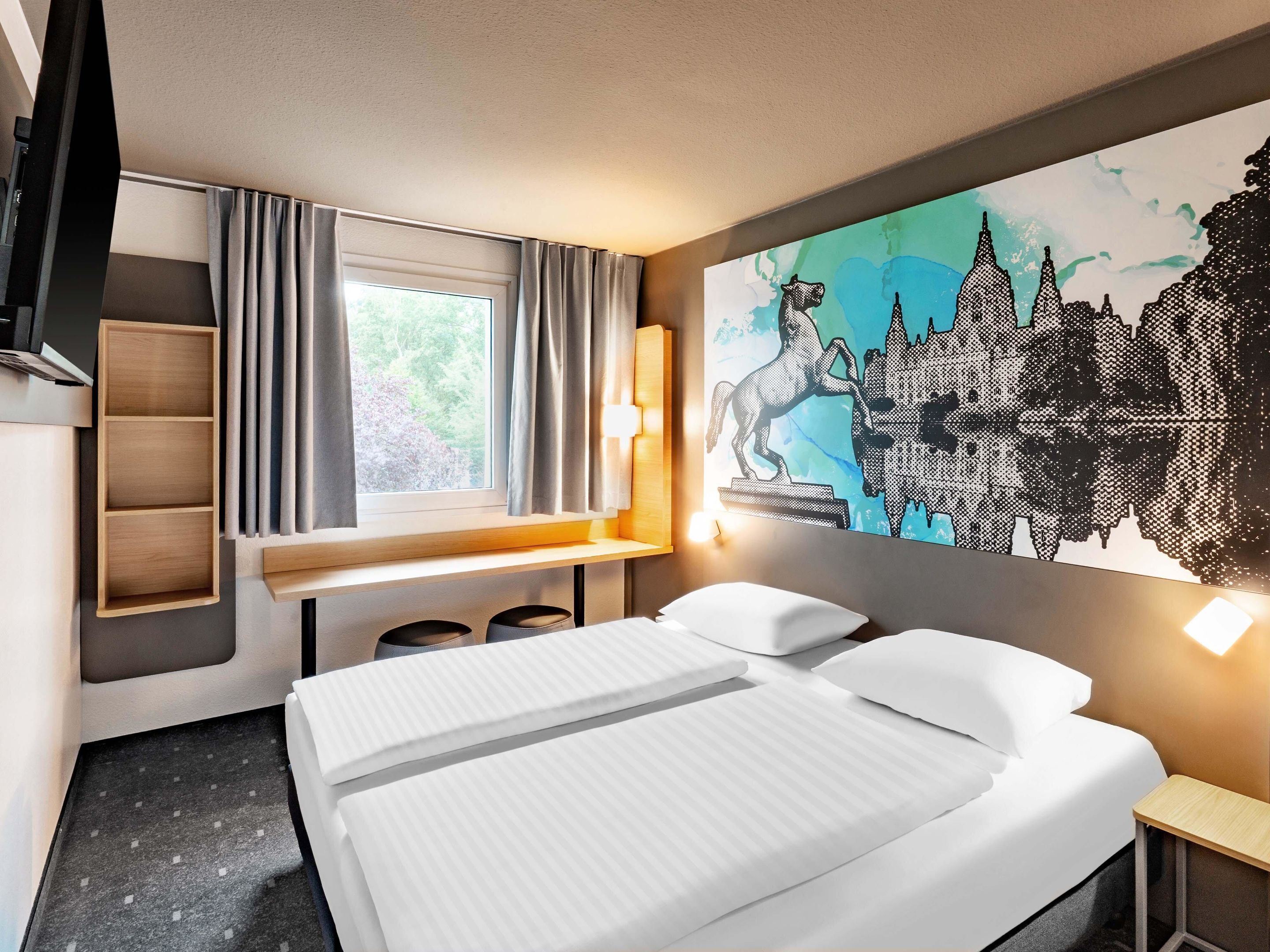 Una habitación de hotel con cama, escritorio, silla, estante y un ventanal con cortinas.