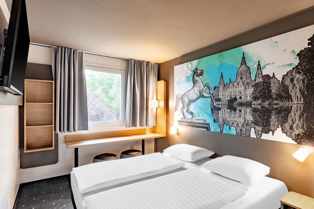 Een hotelkamer met een bed, een bureau, een stoel, een plank en een groot raam met gordijnen.