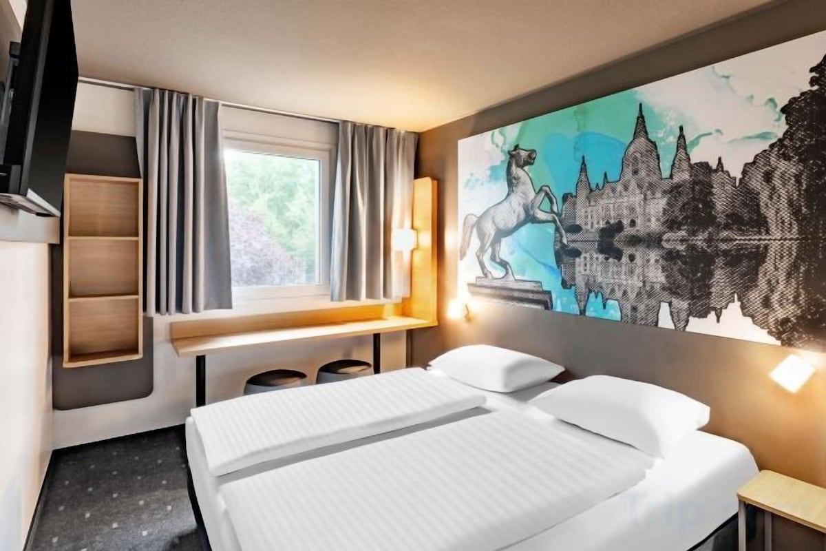Een hotelkamer met een bed, een bureau, een raam met gordijnen, een wandtelevisie en een schilderij van een kasteel.