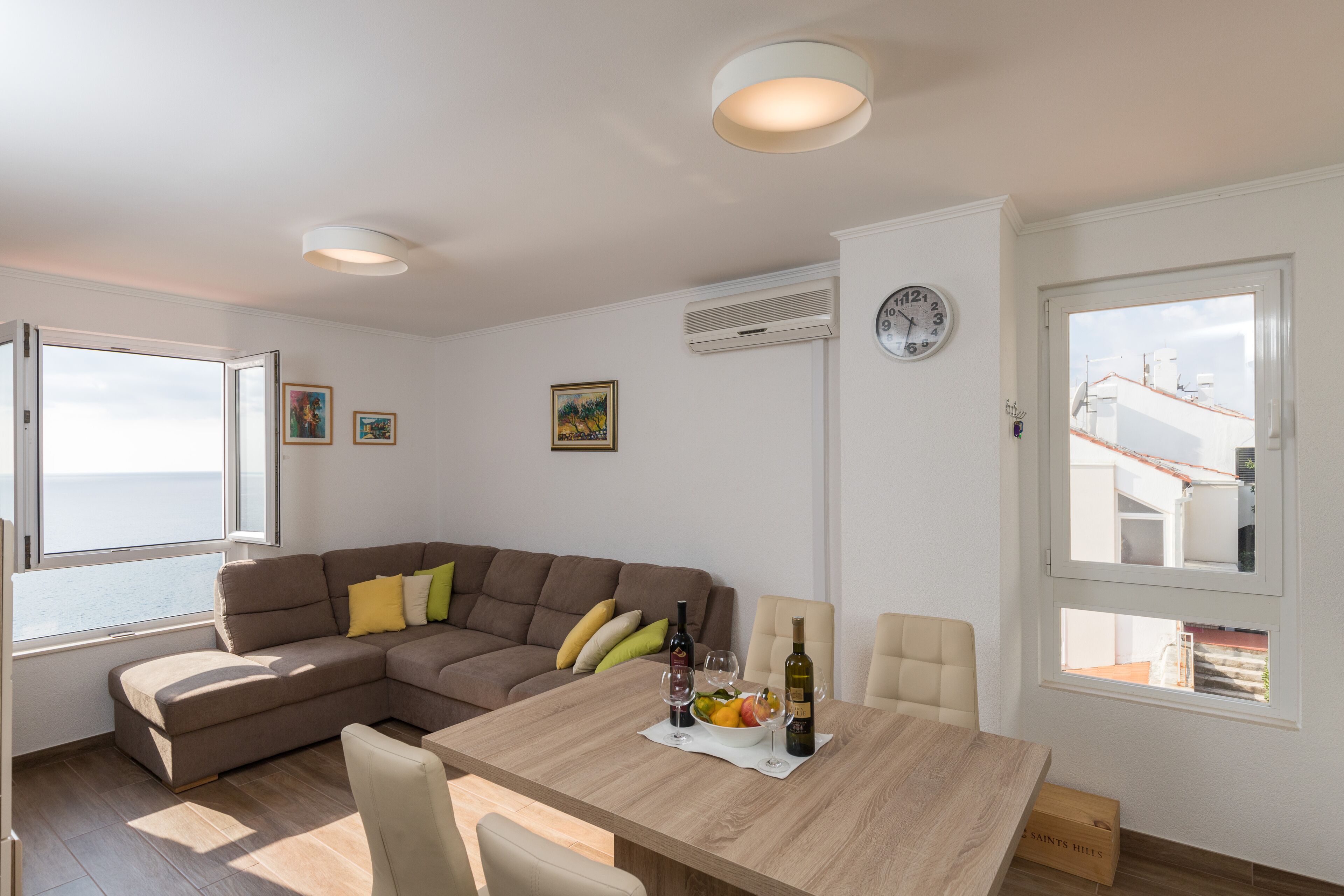 A modern living room with a wooden dining table, a brown sectional sofa, and a view of the sea through a large window.