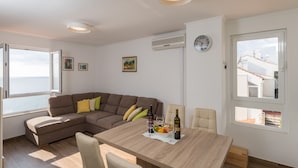 Ein modernes Wohnzimmer mit Esstisch aus Holz, einem braunen Ecksofa und Meerblick durch ein großes Fenster.